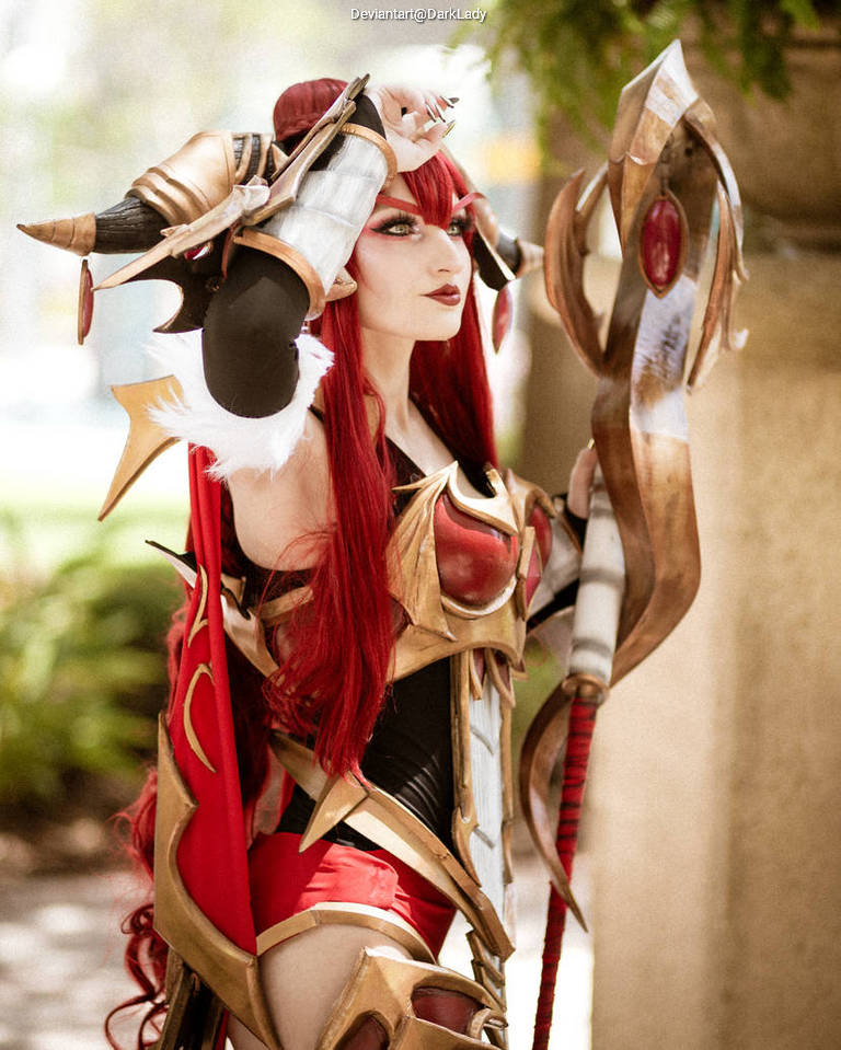 A woman wearing elaborate fantasy armor and a red wig poses confidently, holding a staff adorned with a red gem.