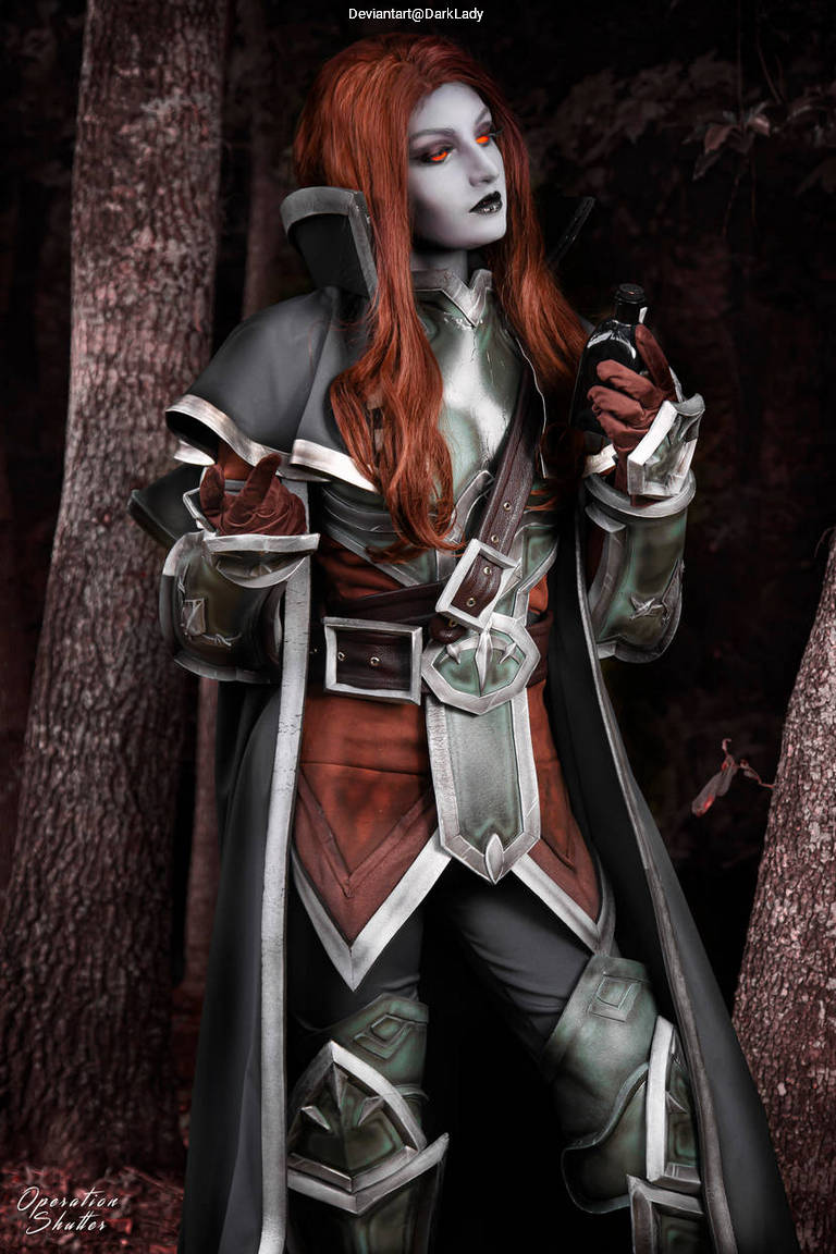 A character in a detailed costume, featuring long red hair and striking makeup, stands against a forest backdrop, holding an item in one hand and dressed in elaborate armor.
