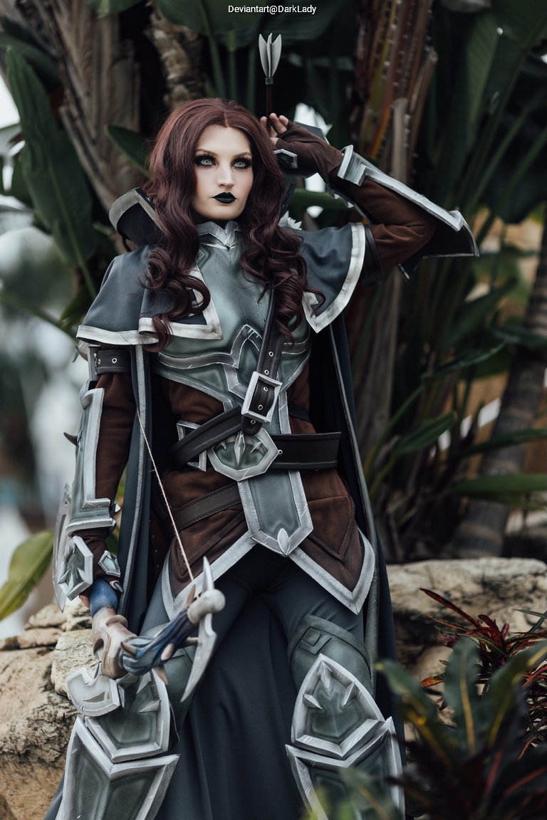 A female cosplayer dressed as a fantasy character, featuring intricate armor with a mix of gray and brown tones, holding a weapon and posing amidst lush greenery.