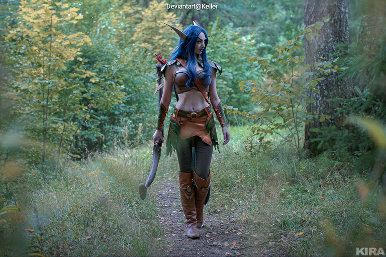 A character in fantasy-themed attire walking along a forest path, surrounded by green foliage and trees.