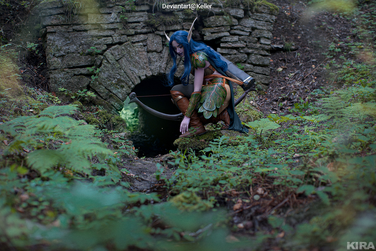 A person dressed in fantasy cosplay attire, featuring greenery and accessories, crouches near a stone archway in a wooded area, holding a bow. Lush foliage surrounds the scene.