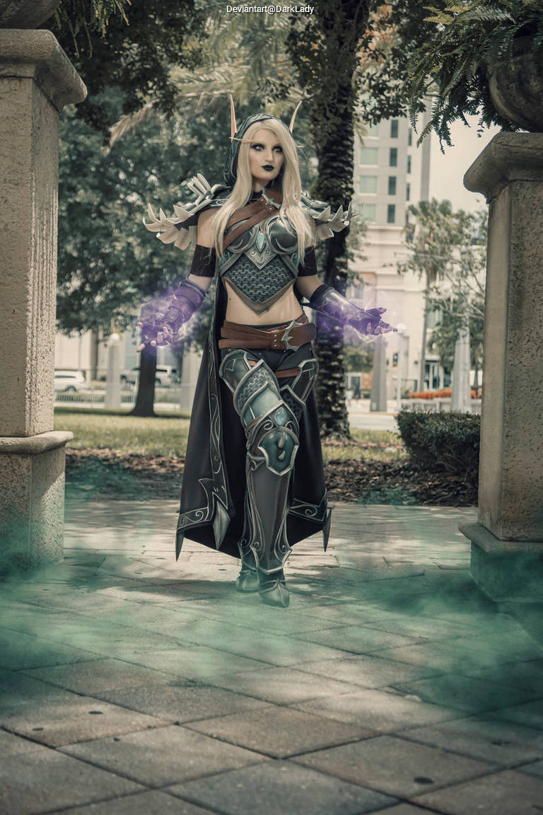 A person dressed in elaborate fantasy cosplay, featuring armor and glowing effects, standing in a park with trees in the background.