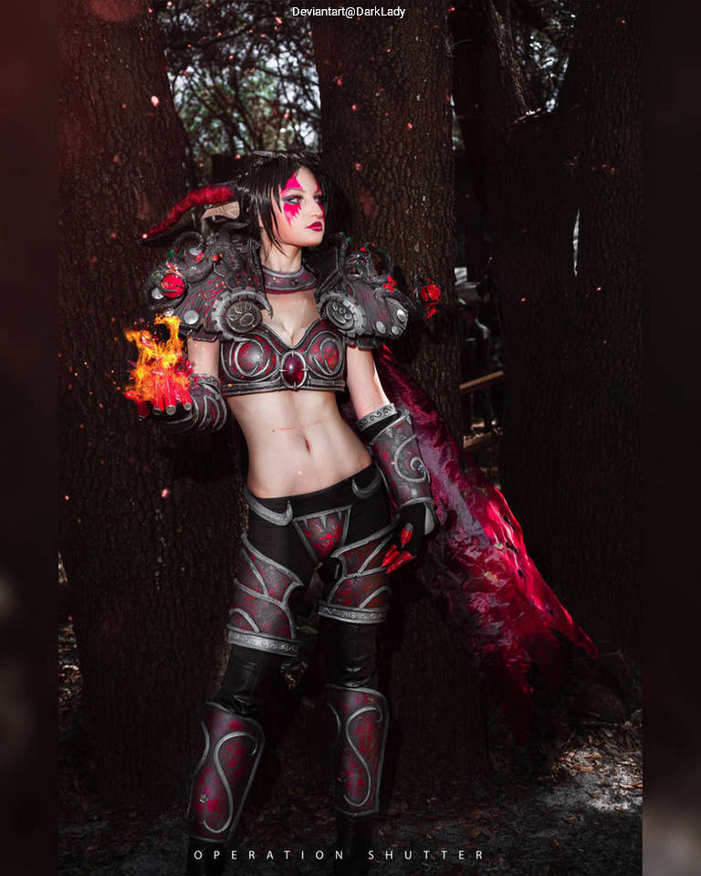 A person in fantasy-themed cosplay, featuring intricate armor with red and black designs, posing against a backdrop of trees. They have colorful makeup and are holding a fiery effect in one hand.