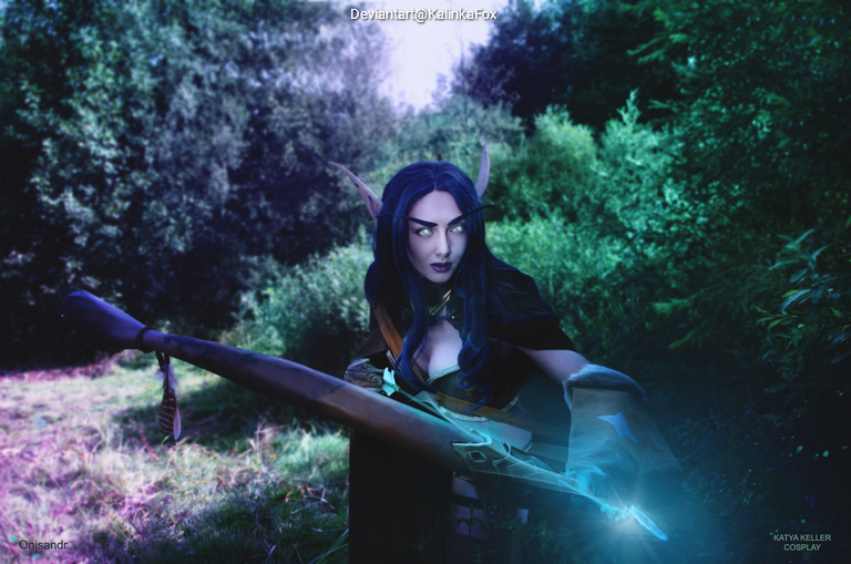 A female cosplayer dressed as a fantasy character, holding a weapon, set against a lush green forest background.