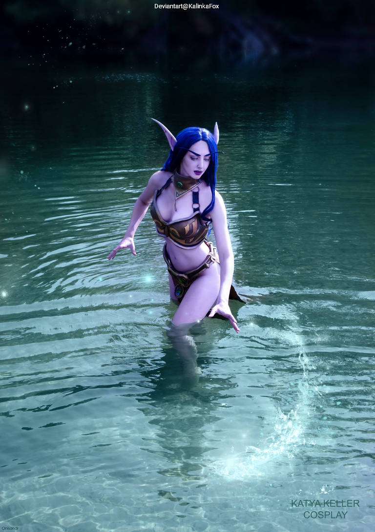 A woman in a fantasy costume wades through a clear, sparkling lake. She has long blue hair and pointed ears, resembling an elf character.