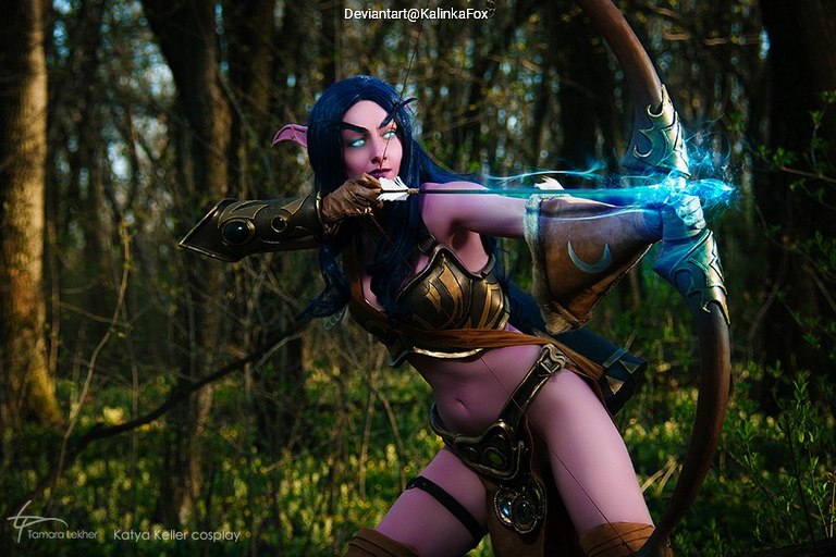 A female cosplayer dressed as a fantasy archer, with blue hair and pointed ears, drawing a bow in a forest setting.