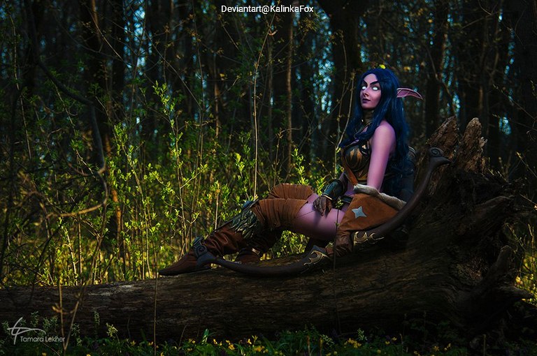 A female cosplayer with blue hair and pointed ears sits on a log in a forest, wearing a fantasy-themed costume with brown and gold accents, surrounded by greenery.