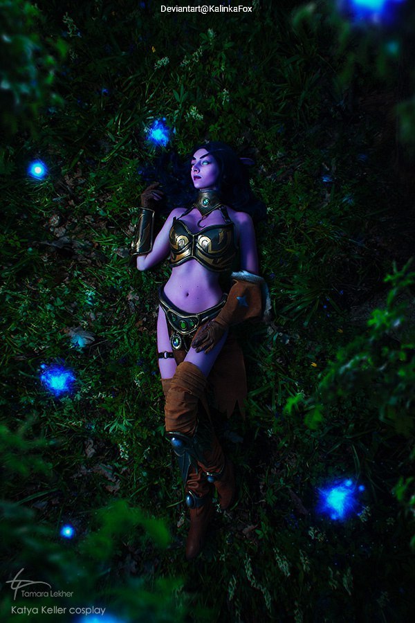 A female cosplayer dressed in a fantasy warrior outfit, lying on grass surrounded by glowing blue orbs.