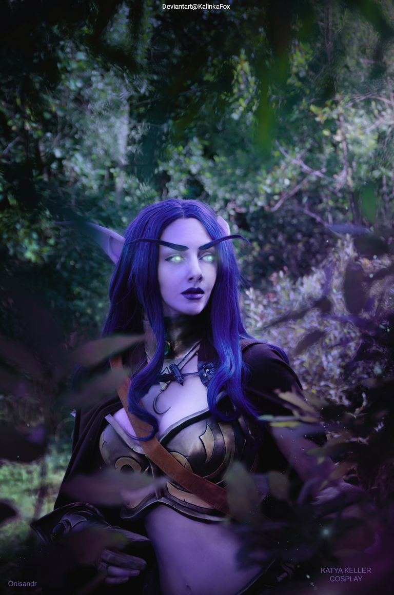 A female cosplayer with long purple hair and glowing eyes, wearing a fantasy costume, stands among lush greenery.