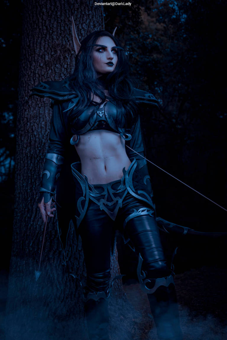 A woman dressed in a fantasy dark elf costume stands confidently in front of a tree, surrounded by a misty atmosphere. Her outfit features dark armor, intricate designs, and she holds a bow, showcasing a fierce and mysterious aesthetic.