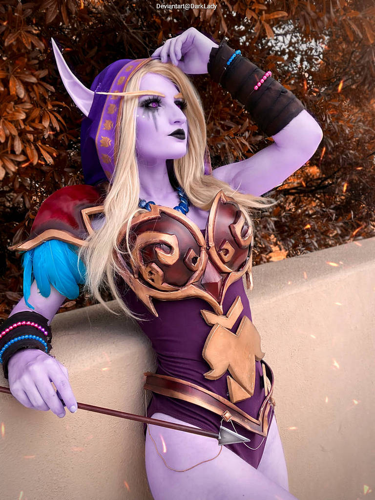 A purple-skinned character in a detailed fantasy costume, posing with a confident expression. The costume features ornate armor with gold accents and a blue shoulder piece, complemented by bracelets and a bow.