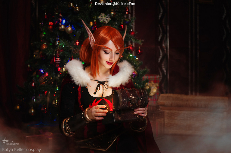 A female cosplayer with red hair and pointed ears is sitting in front of a Christmas tree, holding an ornate book. She is dressed in a dark outfit with white fur accents, creating a fantasy-themed scene.