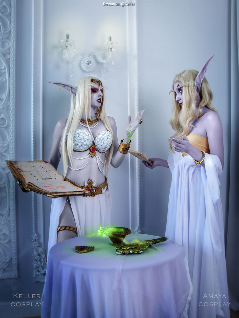 Two cosplayers dressed as fantasy characters, one holding a magical book and the other gesturing towards a glowing object on a table, in an ornate white room.