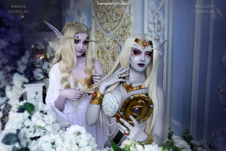 Two individuals in elaborate fantasy character costumes, featuring pointed ears and intricate outfits, surrounded by white flowers and a decorative background.