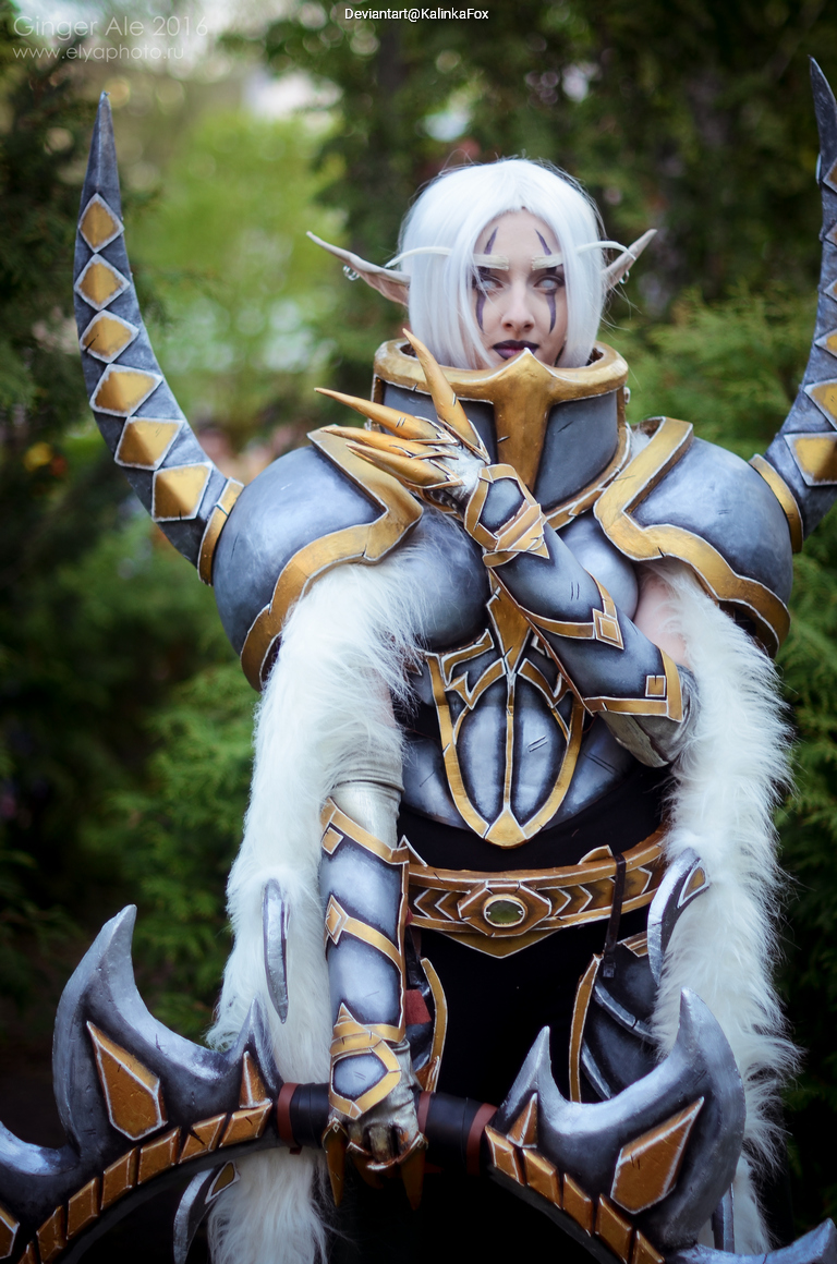 A person in elaborate fantasy armor with silver and gold accents, holding a large weapon, standing outdoors with greenery in the background.