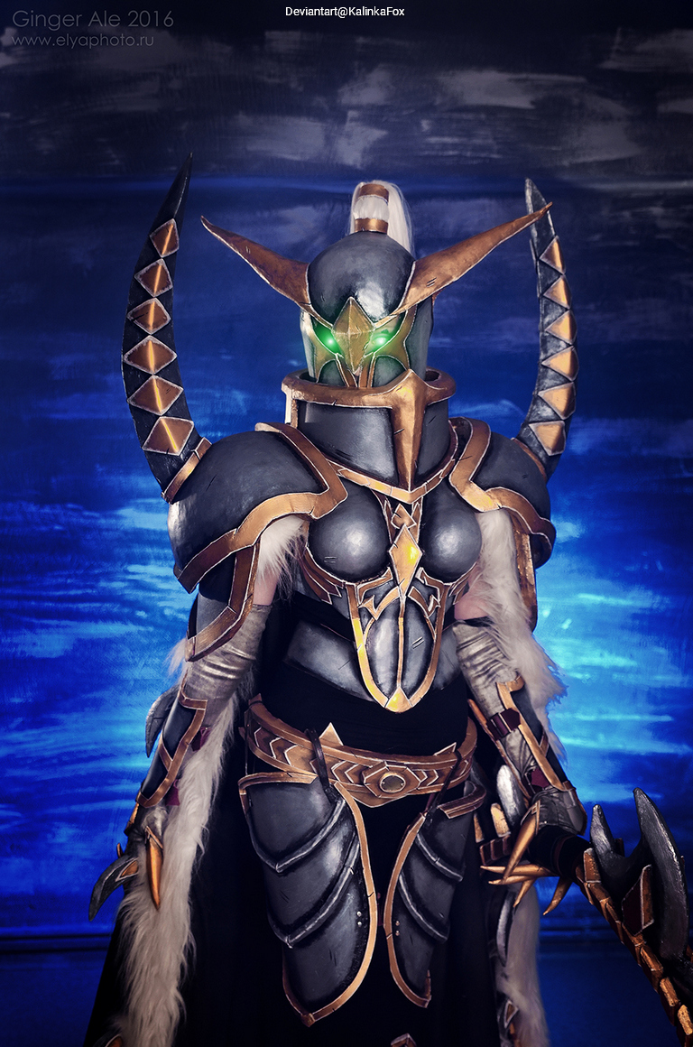 A person dressed in elaborate armor with golden accents and a helmet with glowing green eyes, posing in front of a blue background.