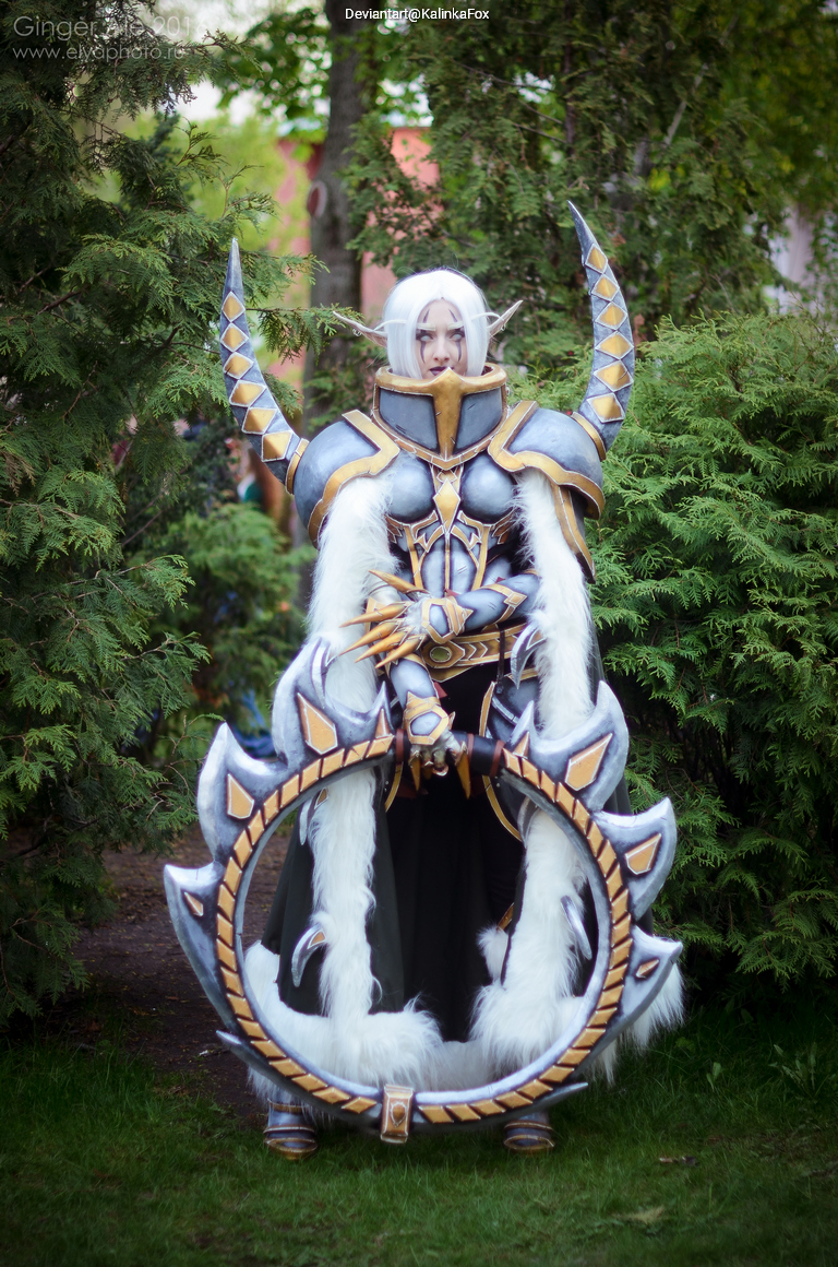 A person in elaborate cosplay, featuring intricate armor with gold and silver detailing, long white hair, and a large circular weapon, stands amid greenery.