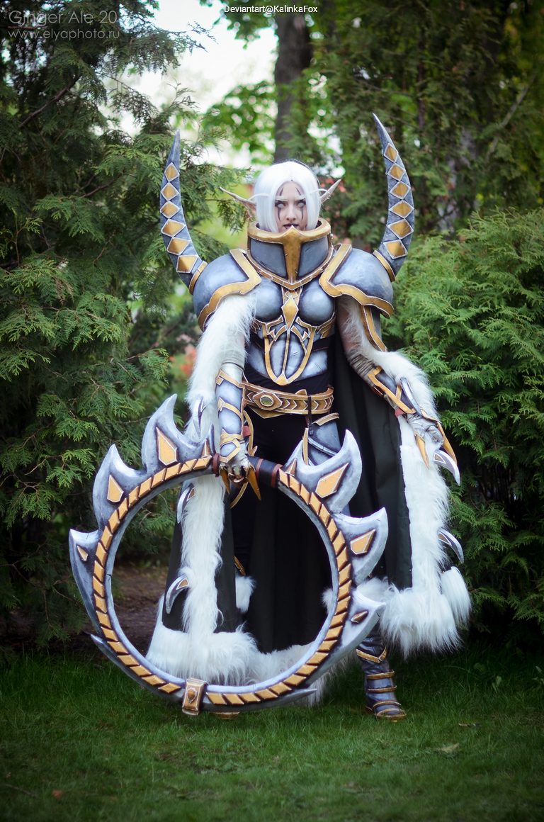 A cosplayer dressed as a fantasy character with long white hair, wearing elaborate armor adorned with silver and gold details. The costume features a flowing cape and a large circular weapon, standing in a natural outdoor setting surrounded by greenery.