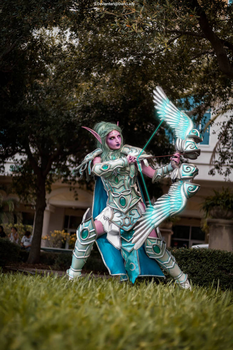 A cosplayer dressed as a fantasy archer with intricate armor and a bow, posed in a grassy area with trees in the background.