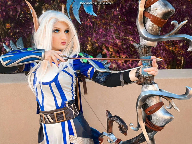 A person in a fantasy-inspired costume, featuring blue and silver armor, is drawing a bow with an arrow, set against a colorful, blurred background.