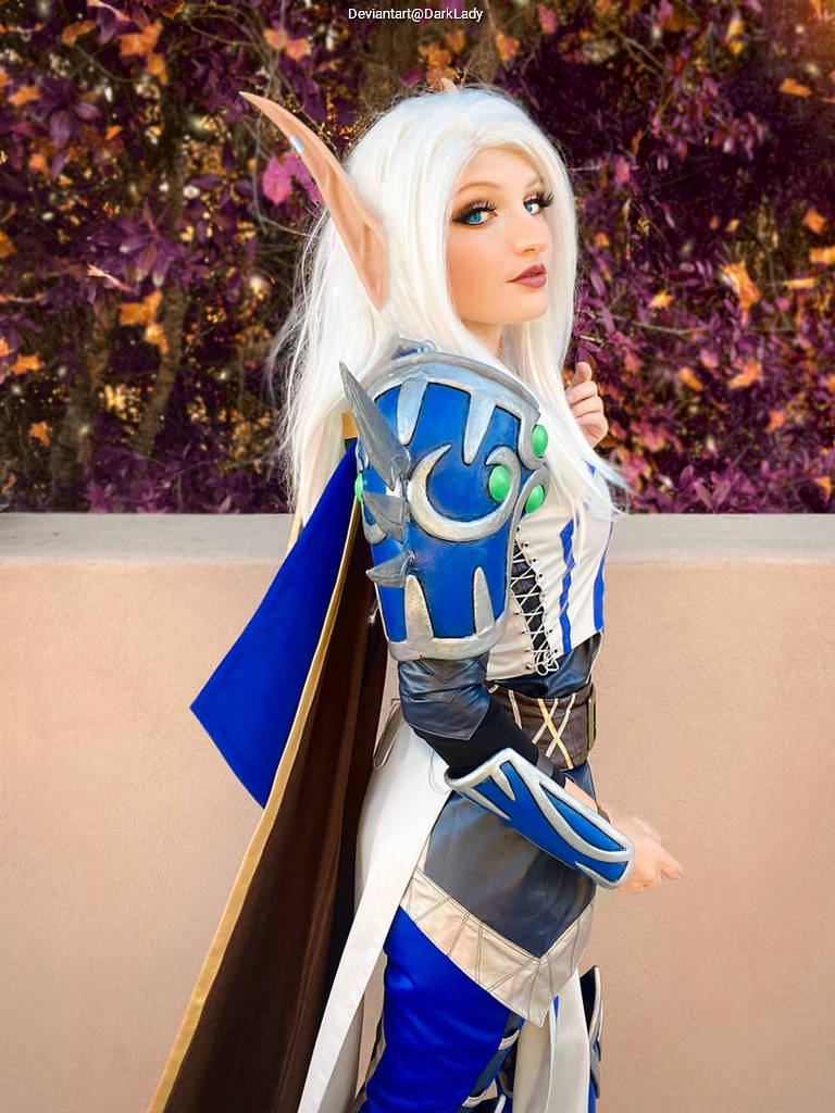 A person in a detailed fantasy costume featuring a blue and silver outfit, long white hair, and pointed ears, posing against a backdrop of purple foliage.