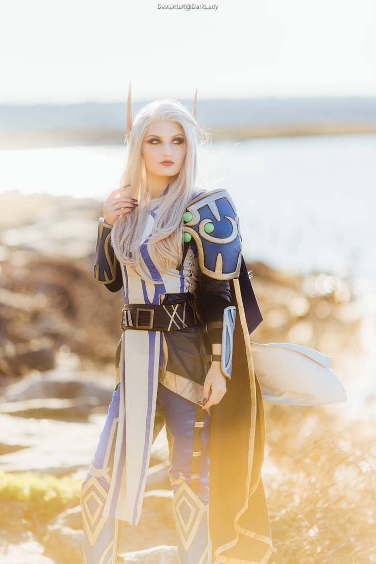 A woman in a fantasy costume with long white hair, pointed ears, and intricate armor stands by a body of water, illuminated by soft sunlight.