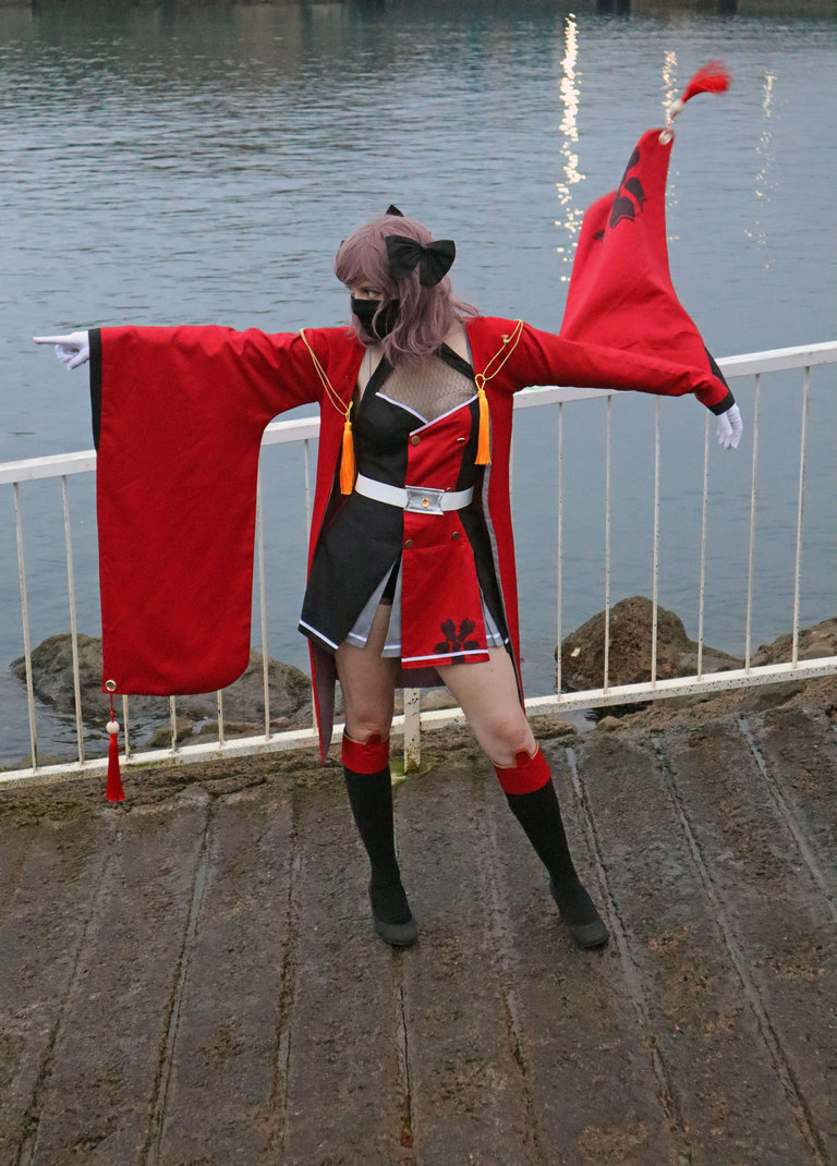 A person dressed in a red and black costume with large sleeves and a mask poses near a body of water, with their arms extended outward.