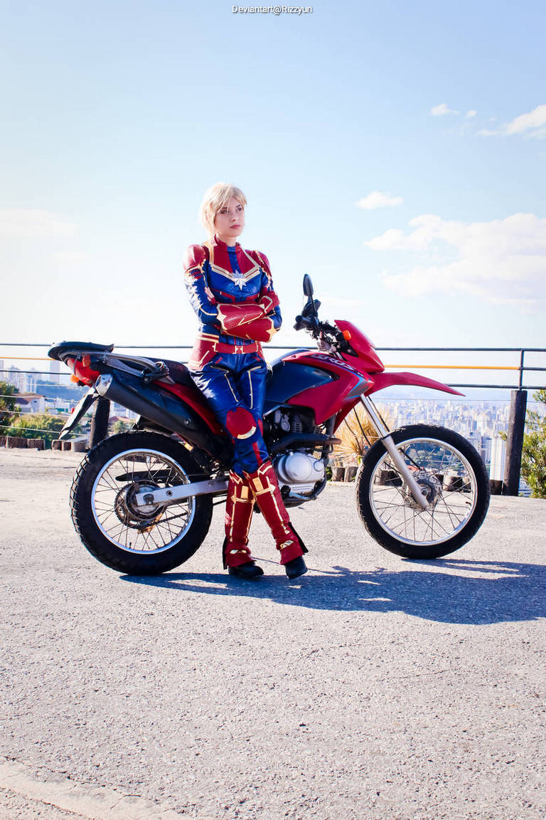 A person dressed in a superhero costume stands confidently next to a red motorcycle. The background features a clear sky and distant cityscape.