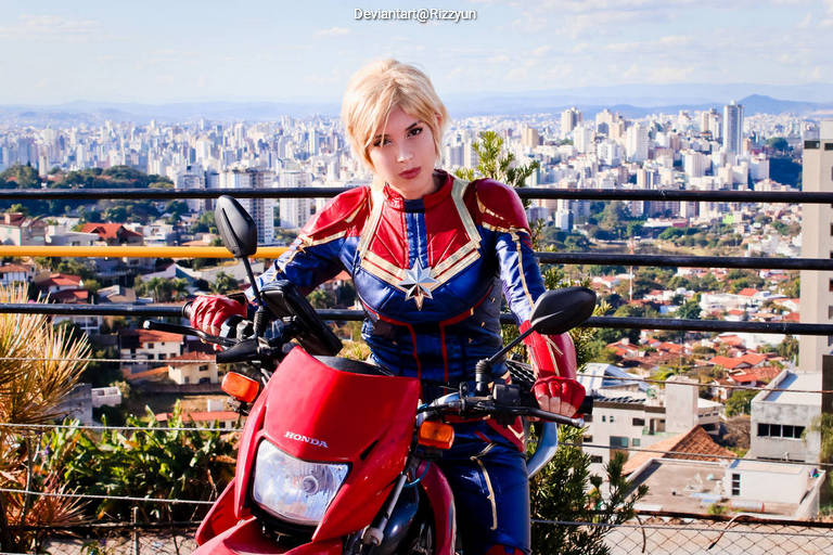 A person dressed as a superhero character poses on a red motorcycle with a city skyline in the background.