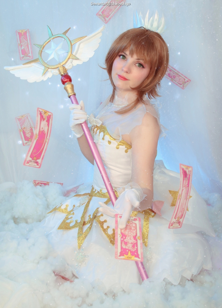 A person dressed in a magical girl outfit holding a staff with a star, surrounded by floating pink cards and a cloud-like background.