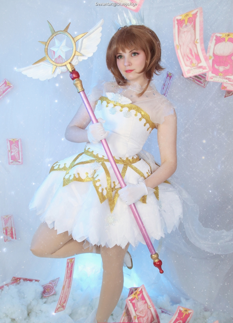 A person dressed in a magical girl costume with a white and gold dress, holding a sparkling staff. The background features a soft, dreamy atmosphere with floating cards.