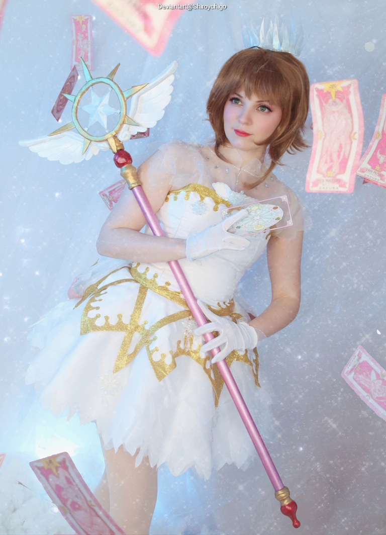 A person dressed as a magical girl, holding a scepter and displaying magical cards, wearing a white and gold costume with a crown and gloves, set against a sparkling background.
