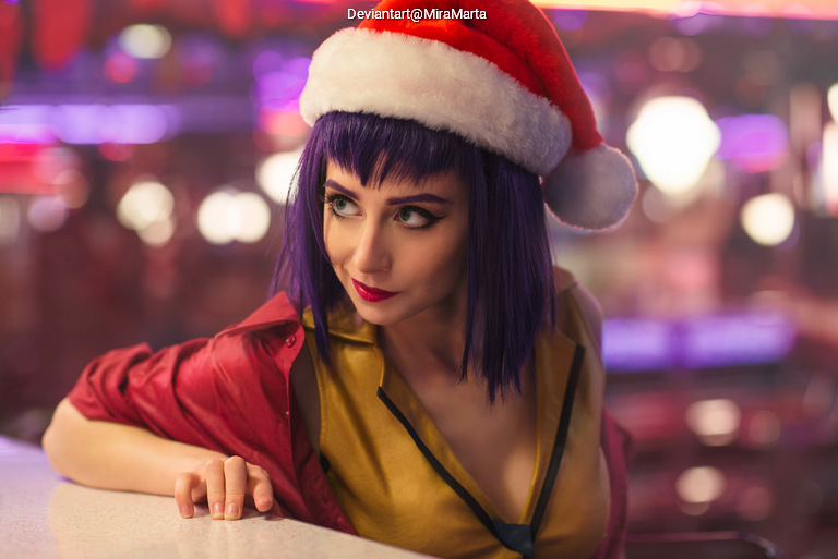 A woman with purple hair wearing a Santa hat and a red jacket, leaning on a counter in a vibrant, colorful setting.