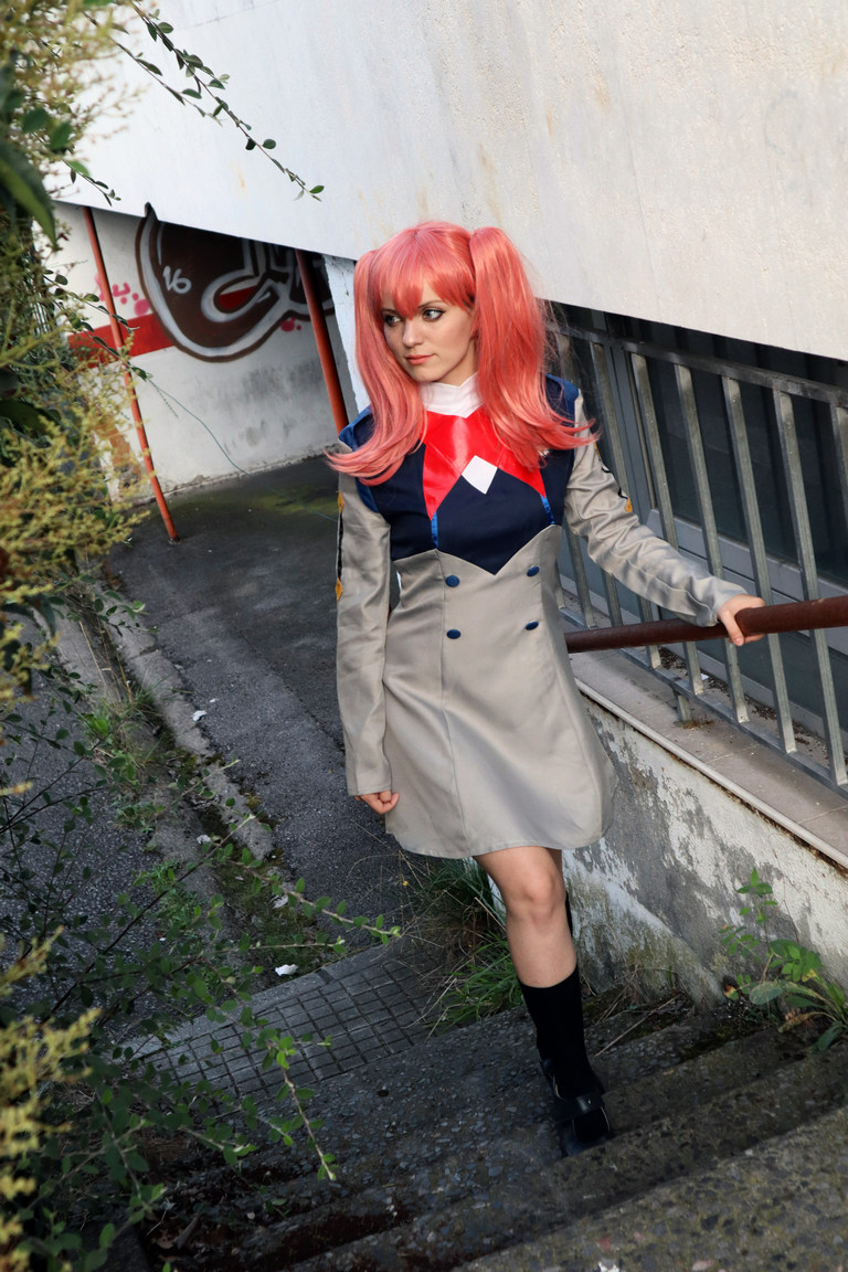 A person with long pink hair styled in pigtails is descending a set of outdoor stairs, wearing a gray and blue coat with a red collar and a short hemline, paired with black knee-high socks and shoes. The background features a wall and some greenery.