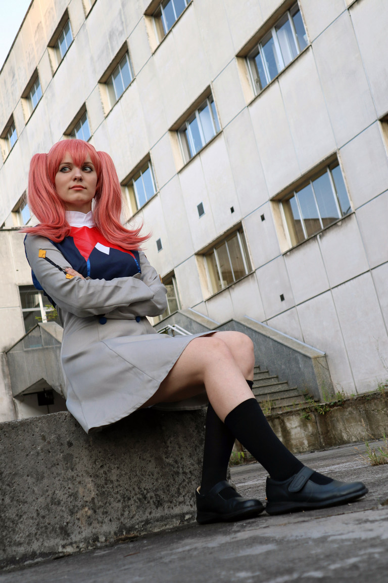 A person in a costume sits confidently on a concrete step, with pink hair and a gray outfit, in front of a building with large windows.