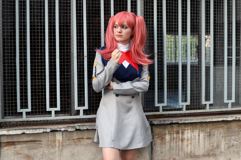A person with pink hair styled in two ponytails wears a gray and blue outfit with a red scarf, standing in front of a grid-like structure.