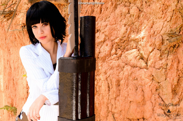 A young woman with short black hair poses with a large sword against a textured orange wall, wearing a white shirt and looking intently at the camera.