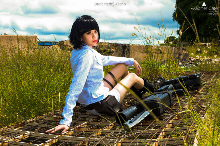 A person with short black hair wearing a blue and white striped shirt and black shorts sits on a metal grid surrounded by tall grass, with a cloudy sky in the background.