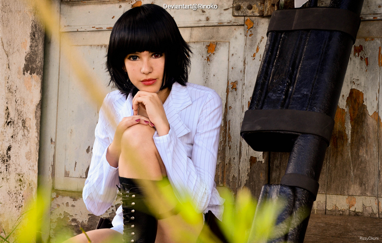 A young woman with a short black bob hairstyle sits against a rustic wall, wearing a white pinstripe blouse and black stockings, with one knee drawn up and a contemplative expression.