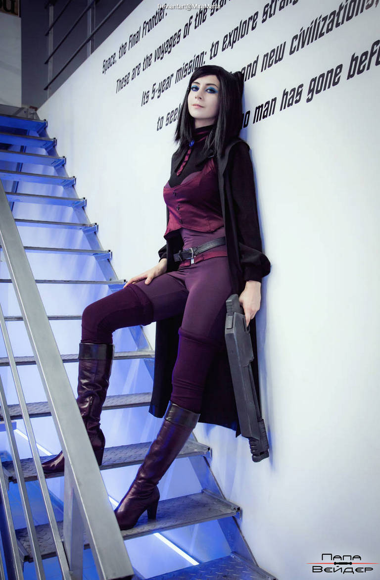 A female cosplayer in a dark, stylish outfit sitting on metal stairs, holding a futuristic gun, with a sci-fi themed background and blue lighting.