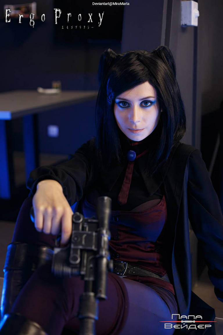 A person in a dark outfit with black hair styled in pigtails, sitting confidently with a weapon resting on their lap. The background features blue lighting and a minimalist setting.