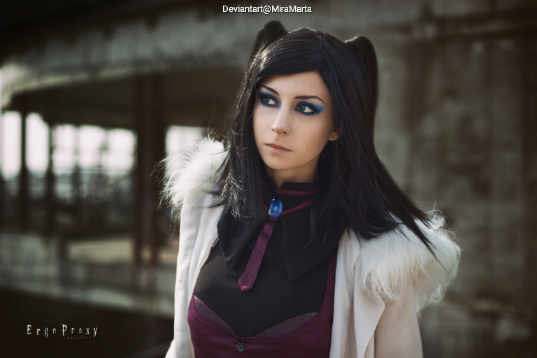 A young woman with long black hair styled in pigtails, wearing a dark outfit with a white collar and a blue gemstone pendant. She has striking blue eye makeup and a serious expression, standing in an industrial setting.