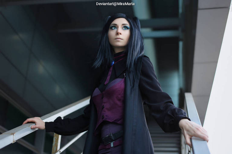 A person with long dark hair wearing a stylish black coat and a purple outfit, posing on a staircase in an urban setting.