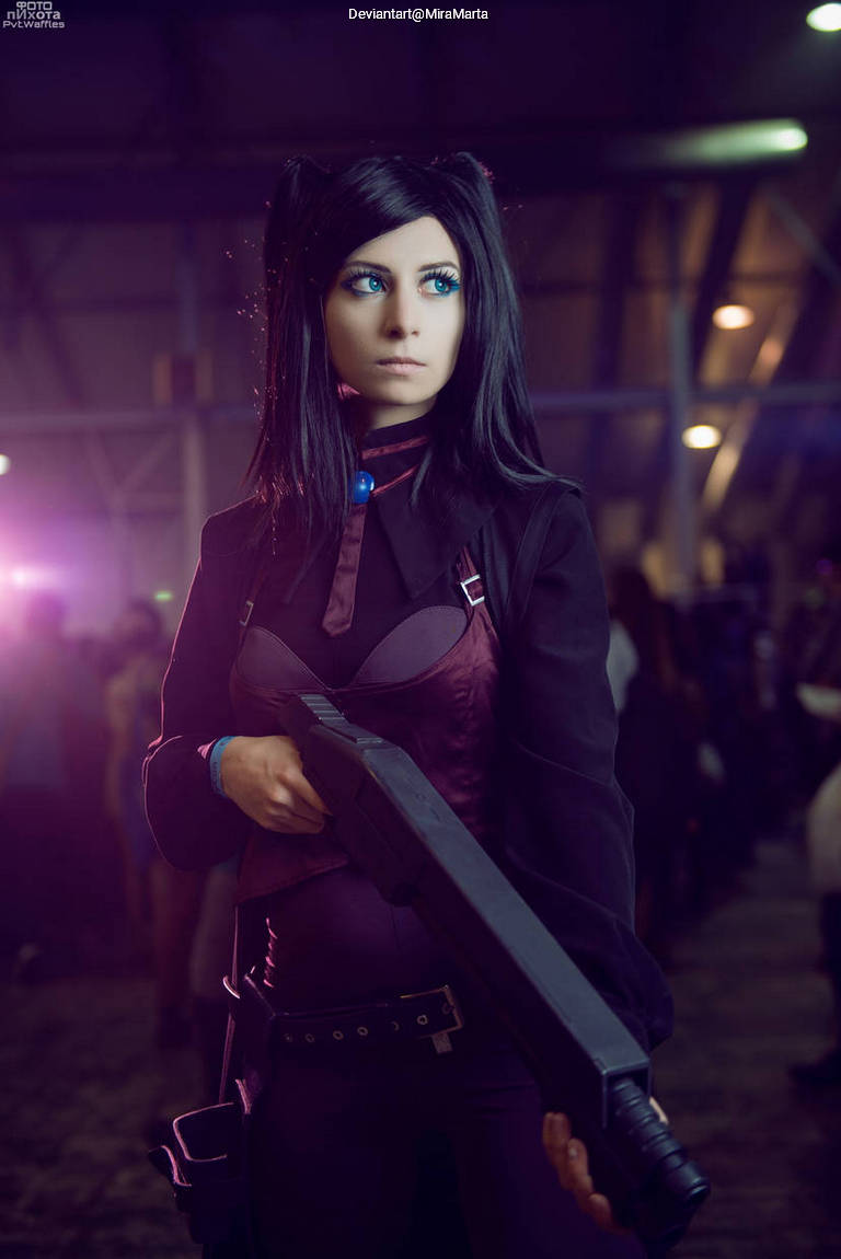 A person in a dark outfit holding a large weapon, with blue eyes and long black hair, posing in a convention setting.