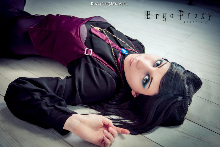 A person lying on the floor dressed in a dark outfit with a purple vest, featuring striking eye makeup and hair spread out around them.