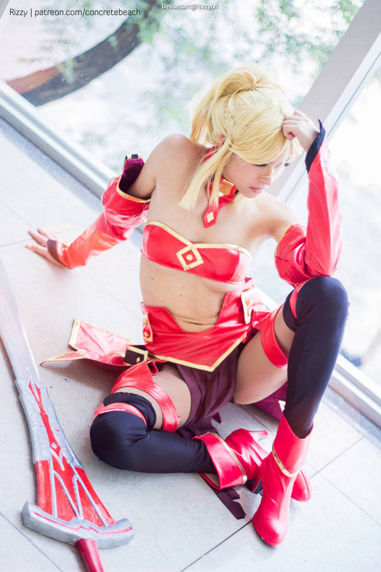 A cosplayer dressed as a character in a red and black outfit, sitting on the floor with a large sword beside her. The setting appears to be indoors with natural light coming through a window.