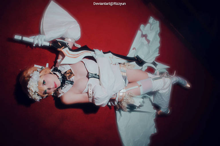A cosplayer dressed as a fantasy character, lying on a red carpet, holding a large weapon, wearing an elaborate white costume with floral accessories in their hair.