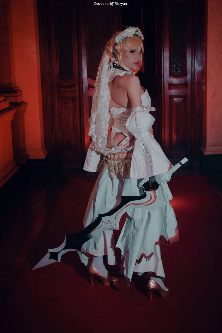 A person dressed in a stylized costume featuring white dress and veil, holding a large, decorative sword, standing in an elegant indoor setting with wooden panels and warm lighting.