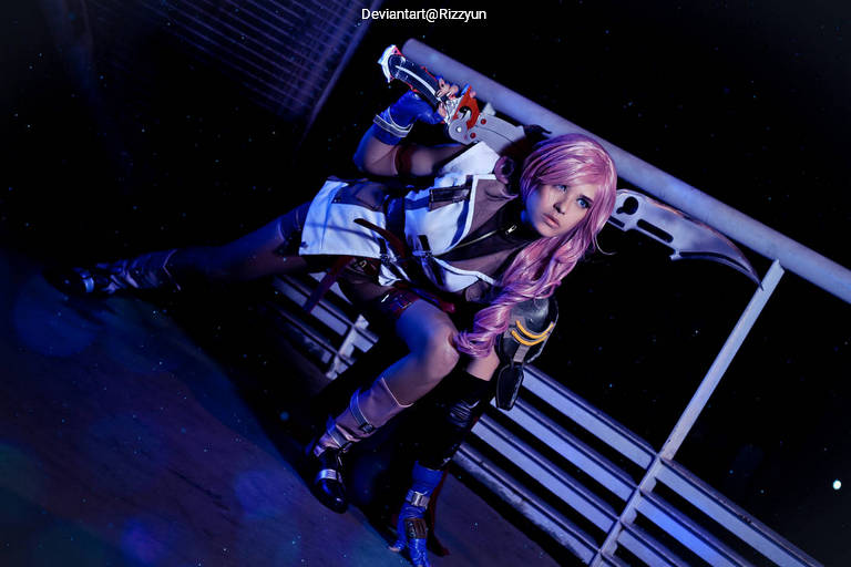 A person in a detailed costume posing in a dynamic stance, holding a large weapon, with pink hair and a strong expression, set against a dimly lit background.
