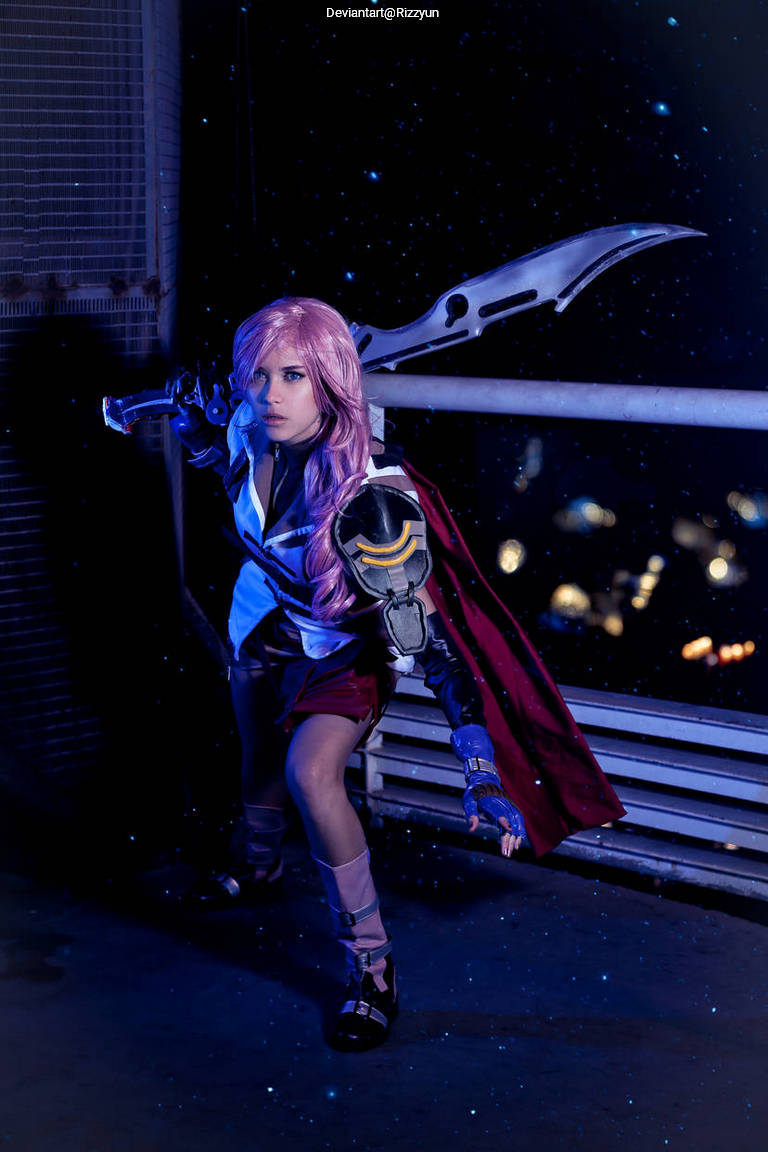 A person dressed in a detailed costume with pink hair, holding a large weapon, posed dramatically against a dark background with city lights.